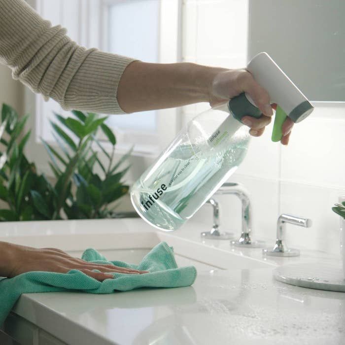 Person cleaning a countertop with a spray bottle labeled "infuse" and a microfiber cloth, surrounded by plants, emphasizing professional cleaning services.