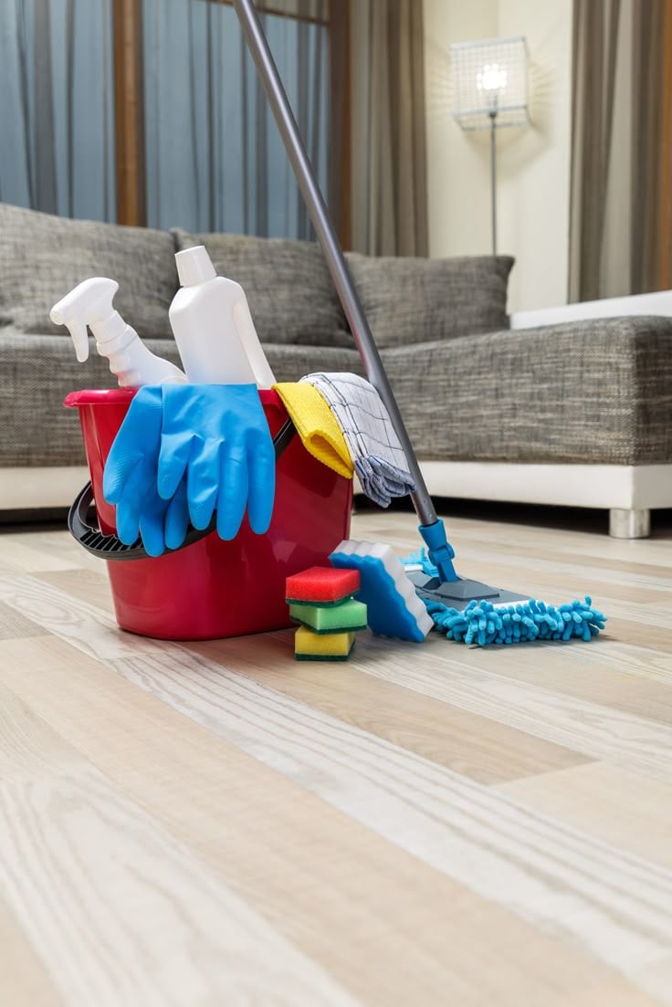 Cleaning supplies and equipment in a red bucket, including spray bottles, sponges, cleaning gloves, and a mop, set on a light wooden floor in a cozy living room environment, emphasizing professional cleaning services.